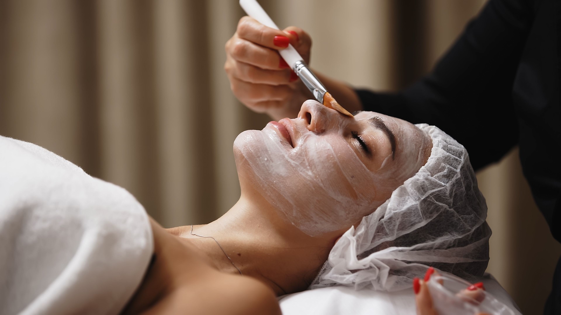 Close-up of a cosmetologist applying a cosmetic peeling mask to a client's face. The scene emphasizes the professional approach to skin care and the relaxing spa experience.